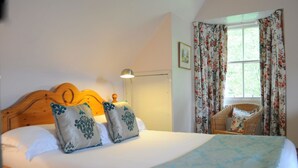 A bedroom with a wooden headboard, two decorative pillows, a bedside lamp, a wicker chair, and a window with floral curtains. - Craigendarroch 2 (Oban)