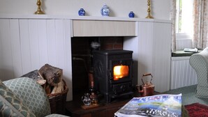 A cozy living room with a wood-burning stove, a wicker basket with logs, a glass pitcher, and a magazine on a coffee table.