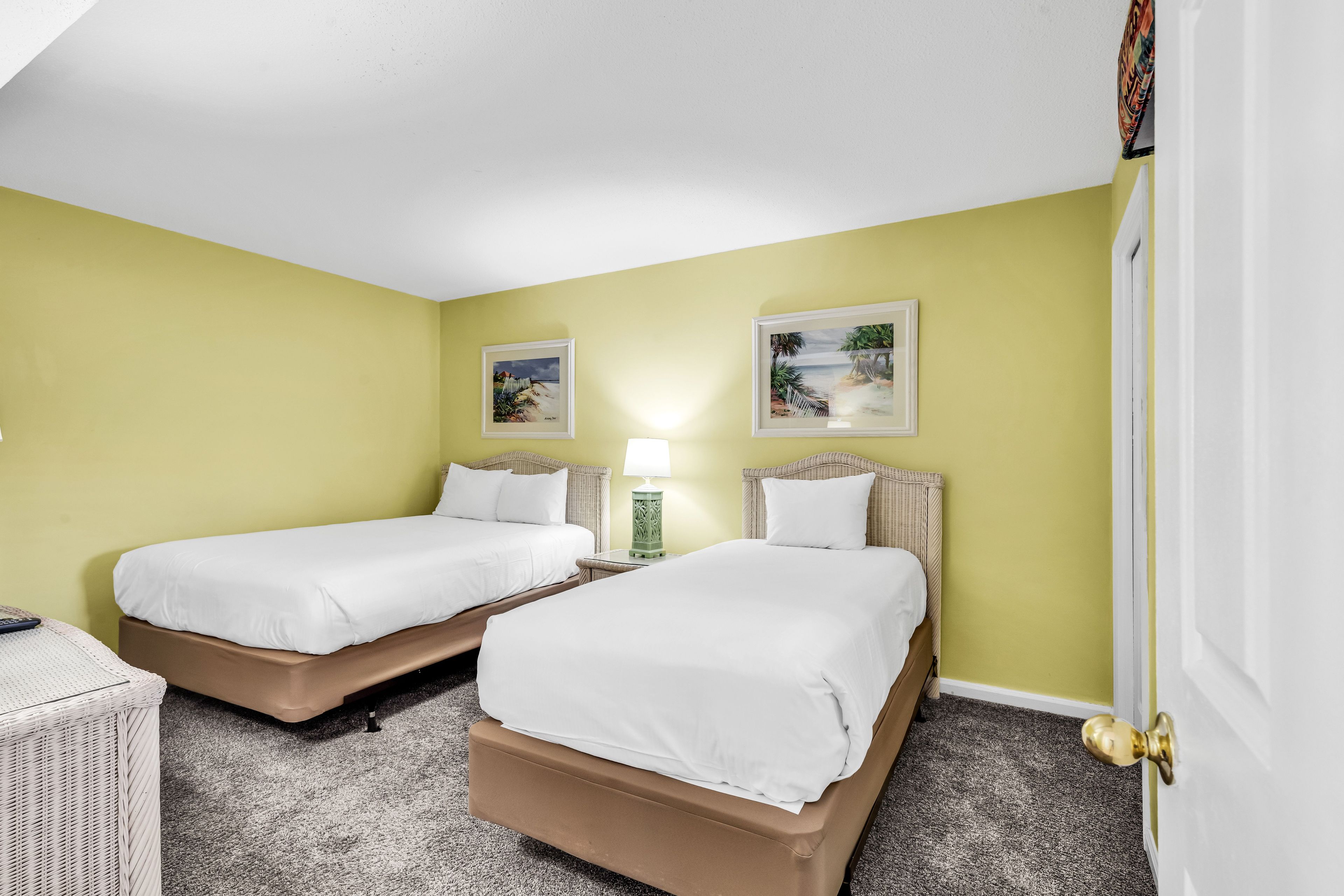 A hotel room with two single beds, a small table, a lamp, and two framed pictures on the wall.
