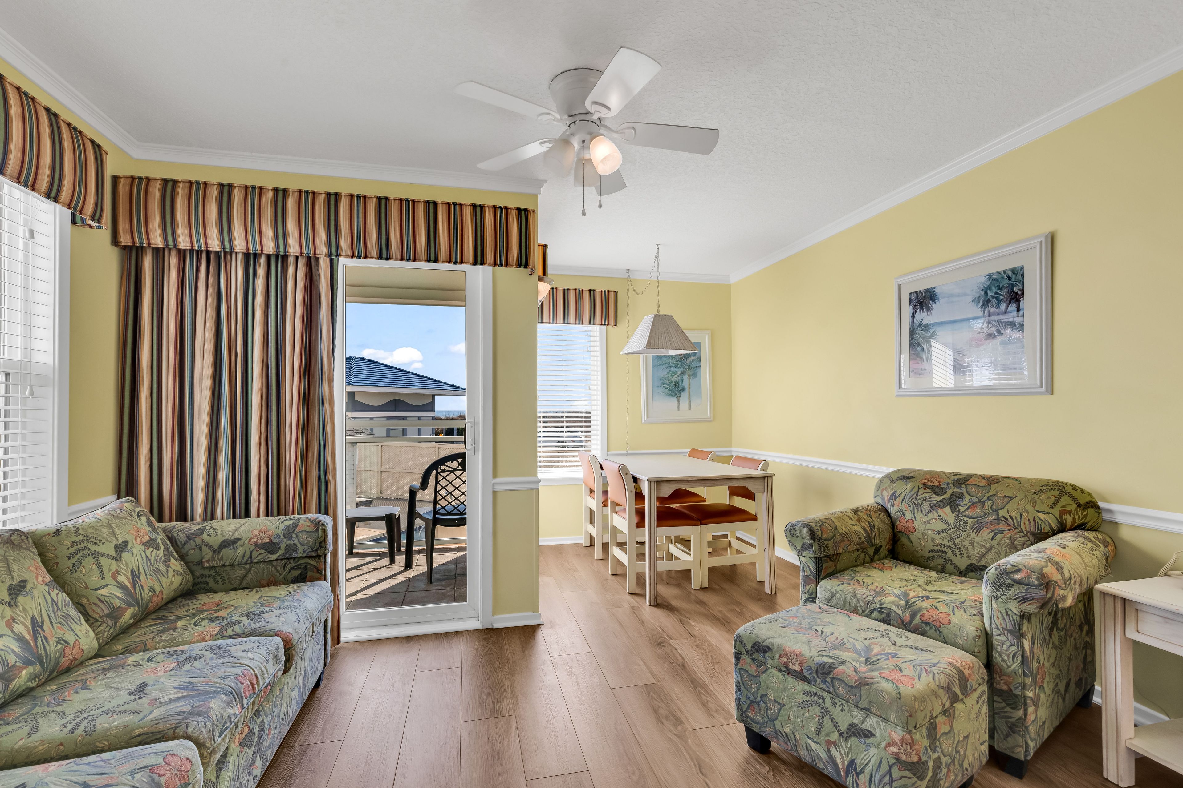 Une pièce comprenant deux canapés à motifs floraux, un ventilateur de plafond, une table à manger avec des chaises, et une vue sur un espace extérieur accessible par une porte vitrée.