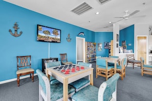 Una habitación con pared azul, muebles de madera, una mesa con un juego de damas y un ventilador de techo.