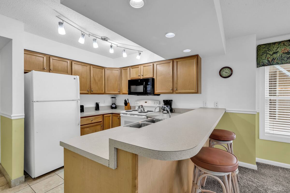 A modern kitchen with wooden cabinets, a white refrigerator, a black microwave, and a large island with stools.