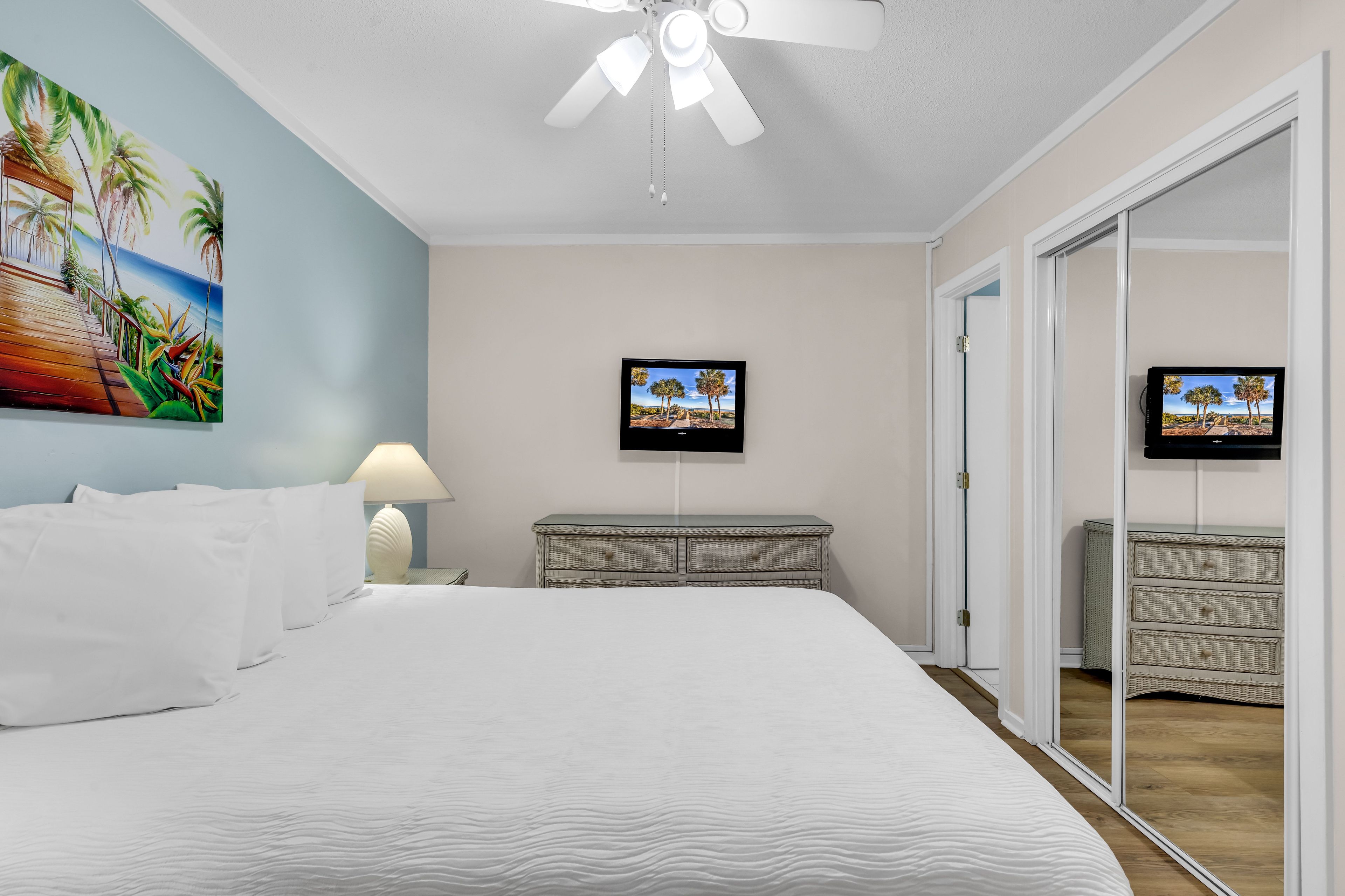 A hotel room with a bed, a ceiling fan, a wall-mounted TV, and a door leading to a bathroom.
