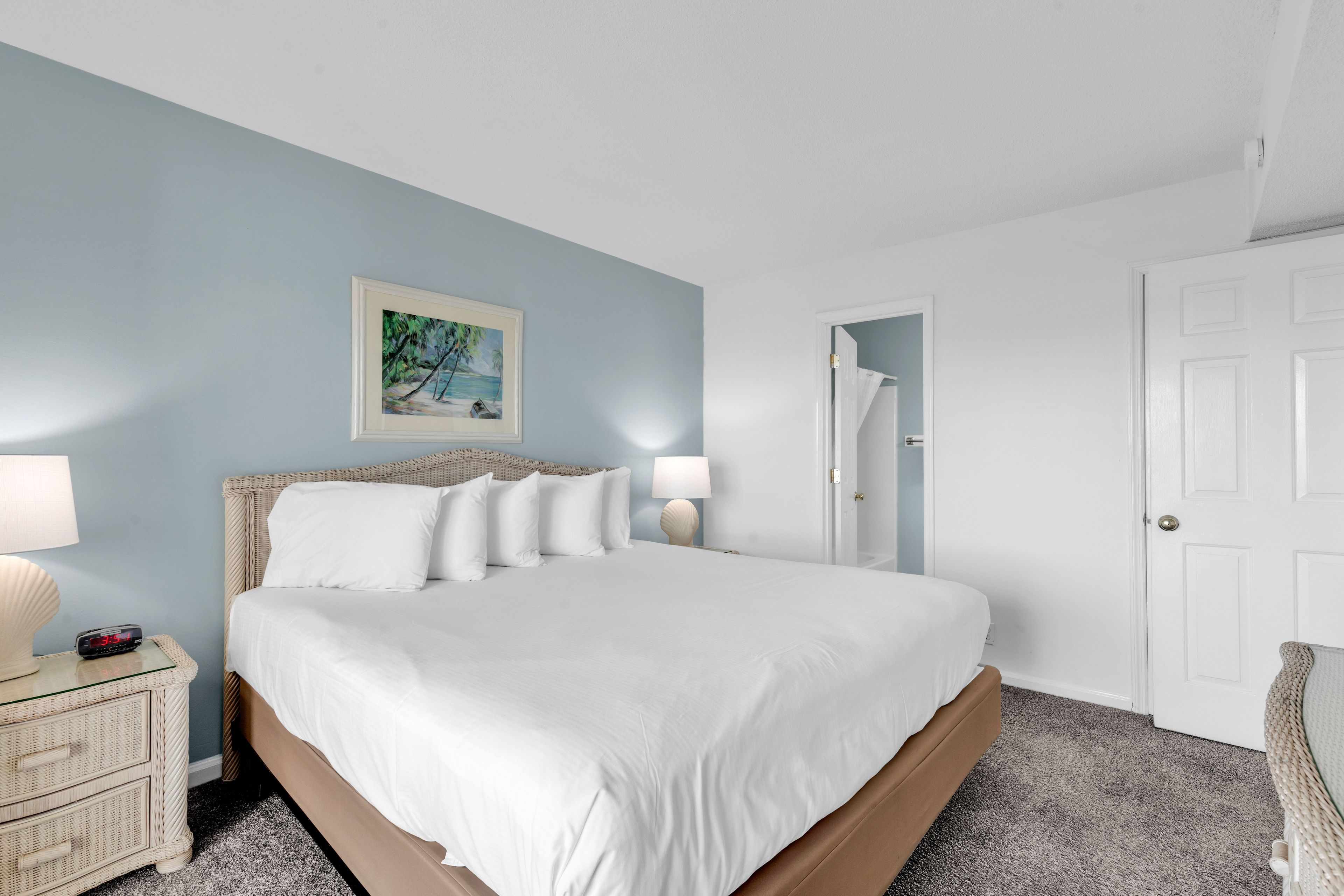 A neatly made bed with white linens, a wicker headboard, and a painting of a tropical scene above it.