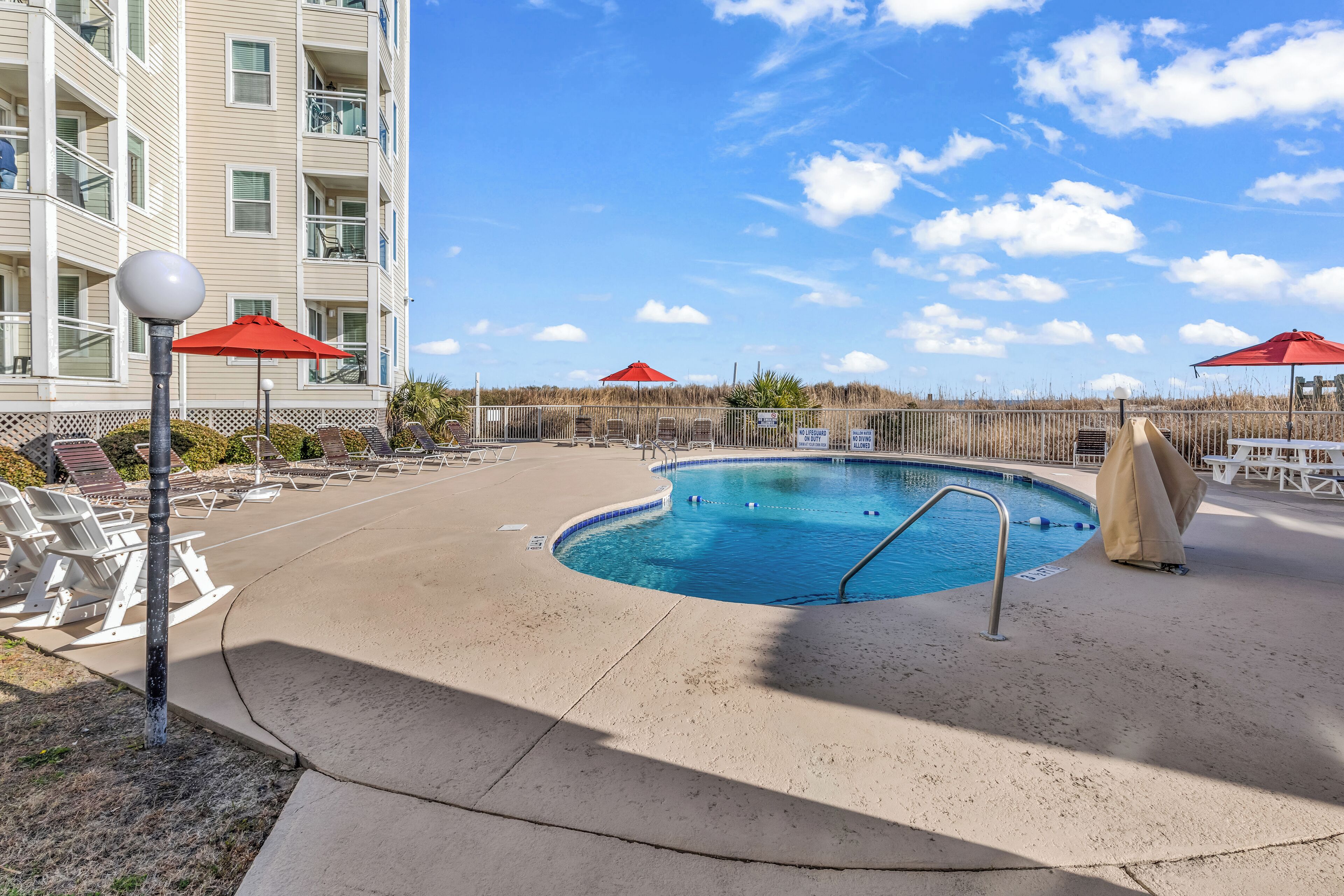 A swimming pool with lounge chairs and umbrellas, surrounded by a fence and a building.