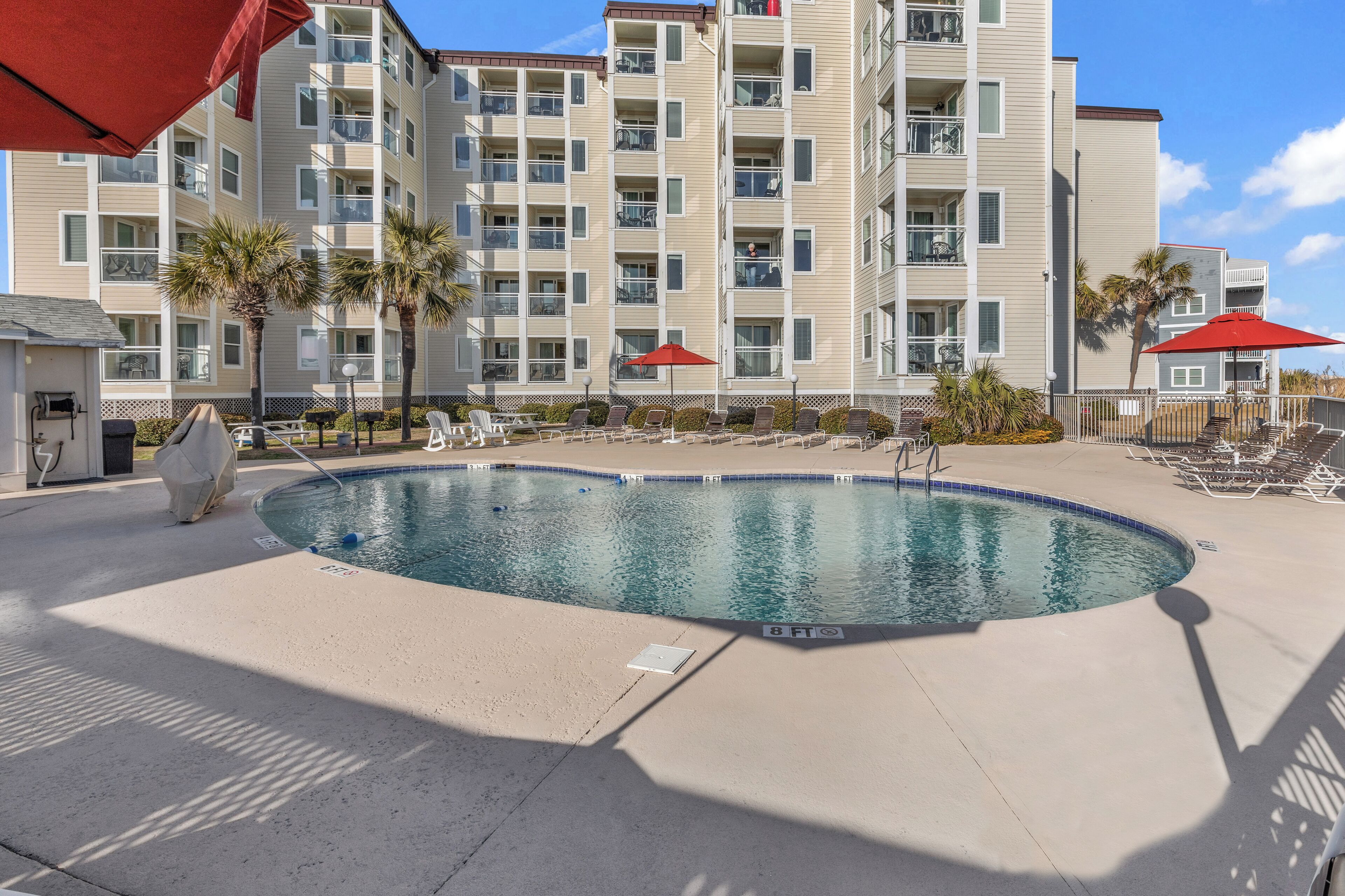 A swimming pool with lounge chairs and umbrellas, surrounded by a multi-story building with balconies.