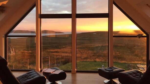 Ein Zimmer mit einem großen Panoramafenster, das Blick auf einen Sonnenuntergang über einer hügeligen Landschaft bietet. Im Raum befinden sich zwei schwarze Stühle und ein kleiner Tisch.