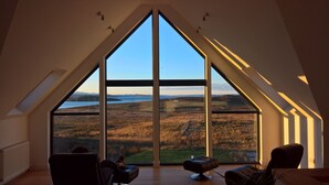 Ein Zimmer mit einem großen, dreieckigen Fenster mit Blick auf eine hügelige Landschaft und ein Gewässer.