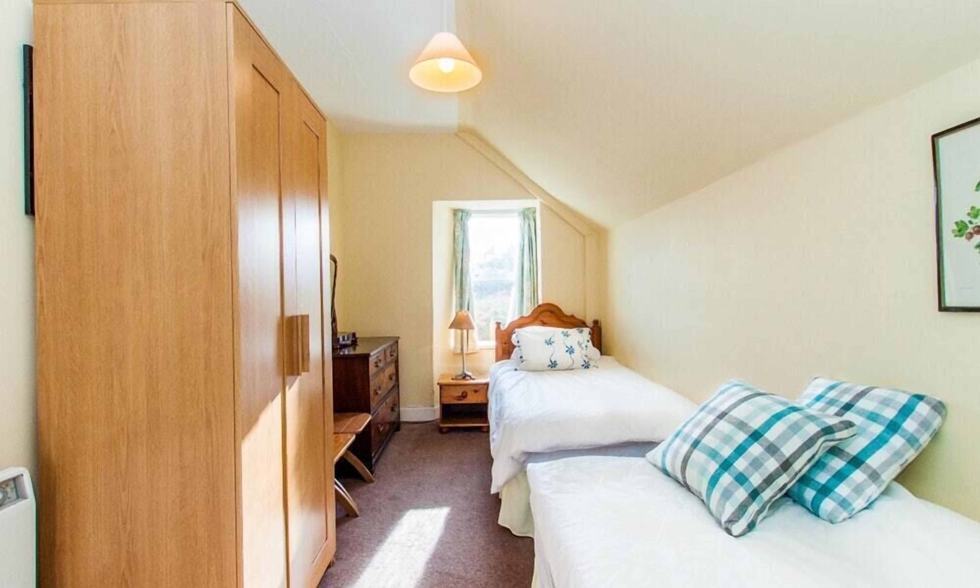 A small, cozy bedroom with a wooden wardrobe, a single bed with a floral pillow, and a nightstand with a lamp.