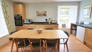 A compact kitchen with wooden cabinets, a microwave, and a dining area with a table and chairs. - Tobaroran (Isle of Colonsay)