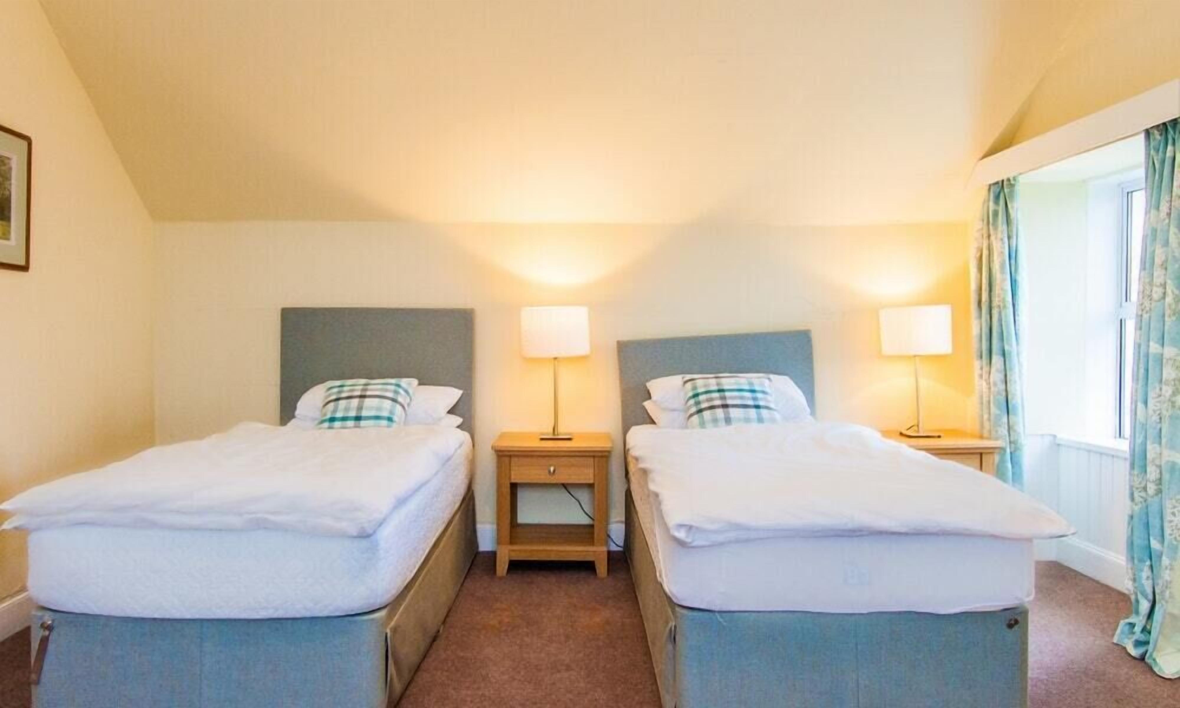 Une chambre d’hôtel avec deux lits simples, chacun doté d’une table de chevet et d’une lampe. La chambre est aménagée de façon simple, avec un sol recouvert de moquette et une fenêtre avec des rideaux.