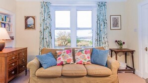 A cozy living room with a beige sofa, floral cushions, a wooden side table, and a bookshelf. - Tobaroran (Isle of Colonsay)