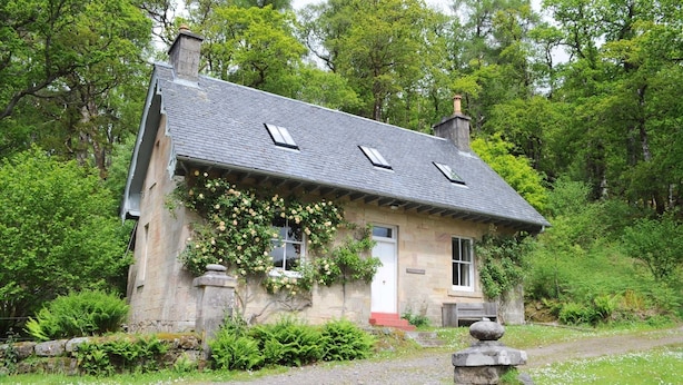 Une maison en pierre avec un toit en pente, un petit porche et un chemin de gravier menant à l’entrée.