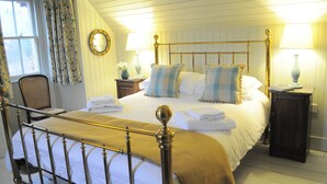 Un lit bien fait, avec une tête de lit dorée, du linge de lit blanc et des oreillers à rayures bleues et blanches. On y trouve deux tables de chevet avec des lampes, une chaise et une fenêtre avec des rideaux.