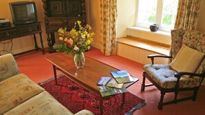 Una sala de estar con una mesa de centro de madera, sillones con estampado floral, un televisor antiguo y una ventana con cortinas.