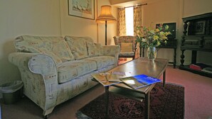 Un salón con un sofá estampado con motivos florales, una mesa de centro con revistas, una lámpara de pie, un jarrón con flores y una estantería.