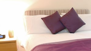 Cama bem arrumada com cabeceira listrada, mesa de cabeceira de madeira com abajur e manta roxa.