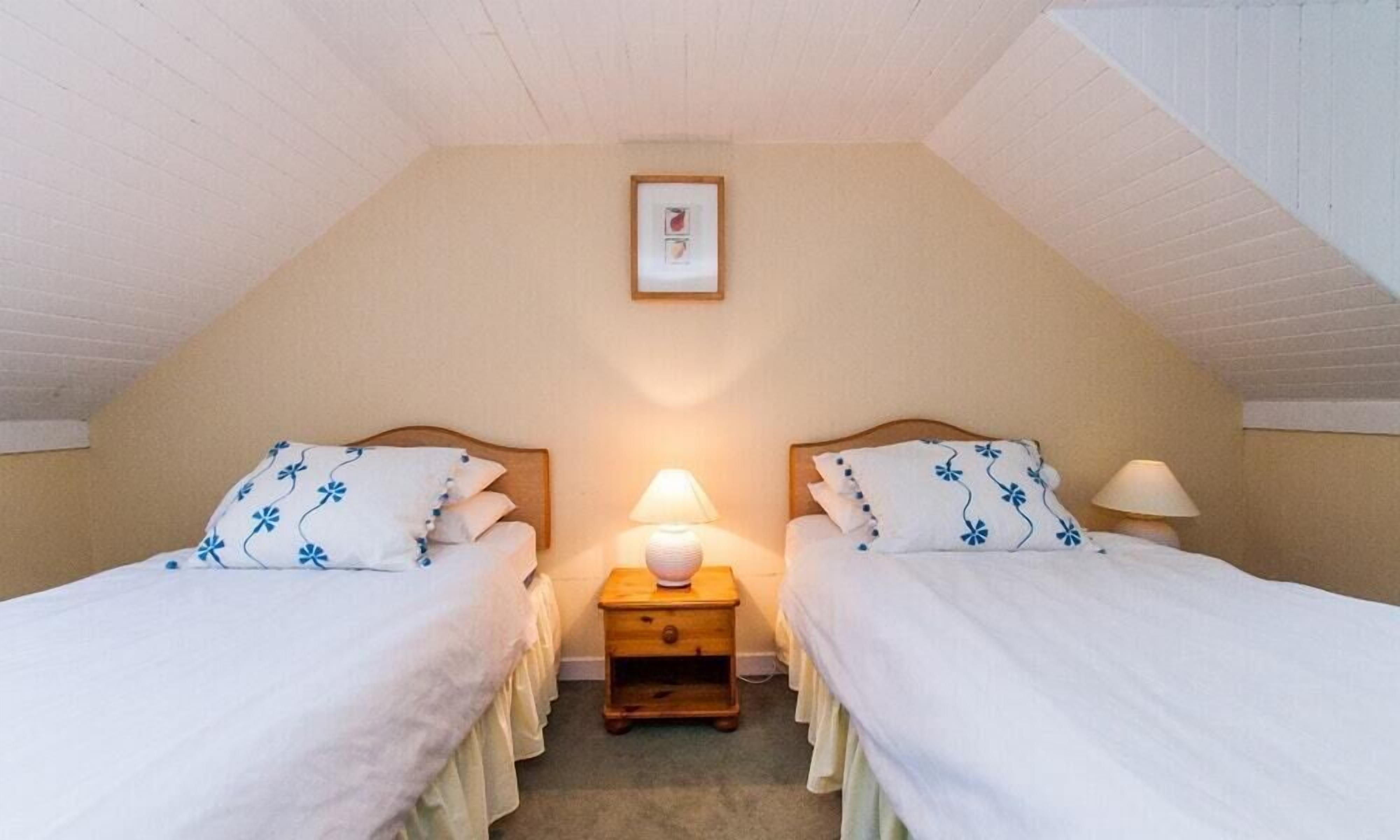 Un dormitorio con dos camas individuales, cada una con cubrecama blanco y almohadas con motivos florales azules, una mesita de noche de madera con una lámpara y un cuadro en la pared.