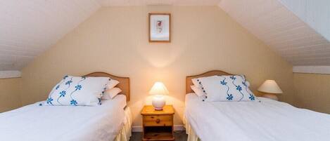 Un dormitorio con dos camas individuales, cada una con cubrecama blanco y almohadas con motivos florales azules, una mesita de noche de madera con una lámpara y un cuadro en la pared.