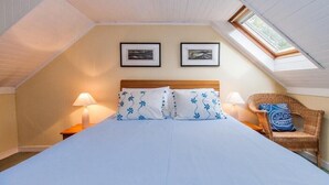 A bedroom with a sloped ceiling, a bed with white bedding and blue floral pillows, two framed pictures on the wall, a wicker chair with a blue pillow, and two bedside lamps.
