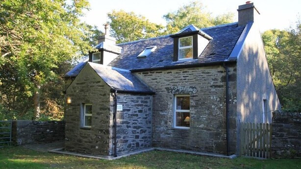A stone house with a sloped roof, two windows, and a small attached structure.
