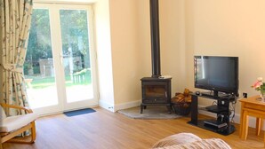 A cozy living room with a wooden floor, a sofa, a television, a fireplace, and a sliding glass door leading to a garden.