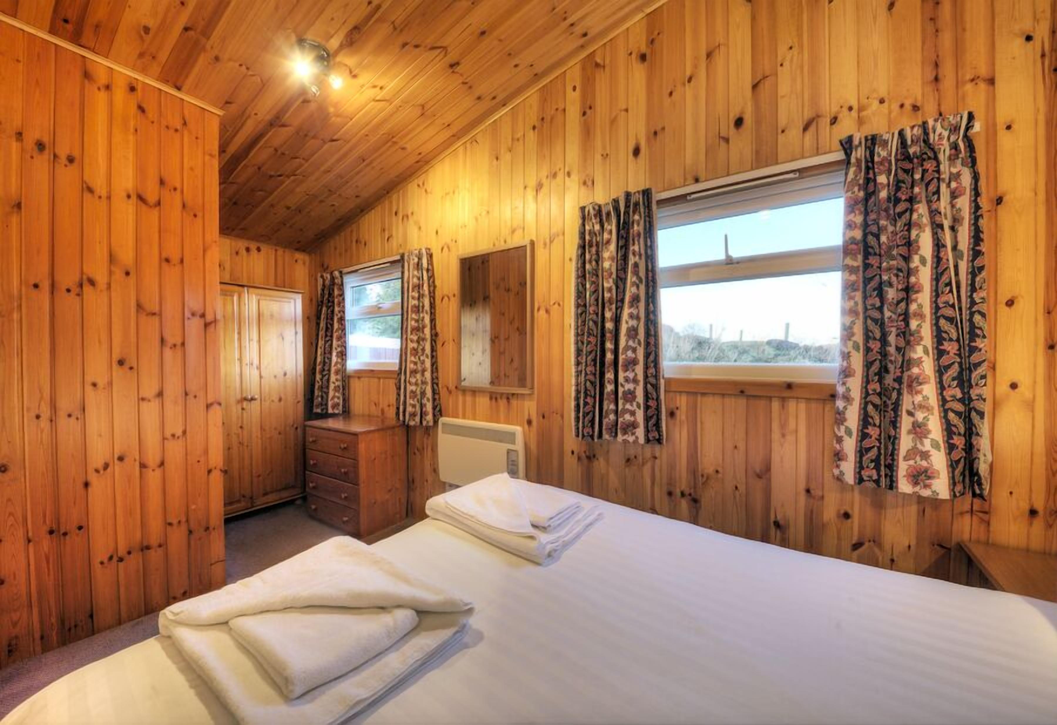 Une chambre à coucher aux murs lambrissés, avec un lit, une commode et une fenêtre ornée de rideaux à motifs floraux.