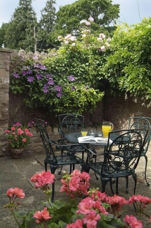 A patio with a table and chairs, surrounded by flowering plants and a stone wall.