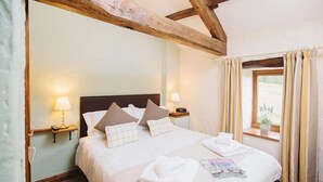 A hotel room with a large bed, two bedside lamps, a window with curtains, and a wooden ceiling beam. - Gelt Cottage (Hot Tub) (Brampton)