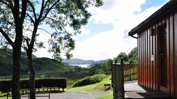 A wooden cabin with a porch, surrounded by a grassy area and trees, overlooking a valley with a lake and hills in the distance. - Ash lodge (Oban)