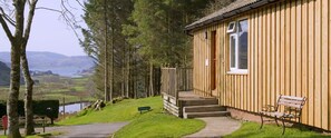 Eine Holzhütte mit einer Holzterrasse und einer Bank, umgeben von Bäumen und Blick auf ein Tal.