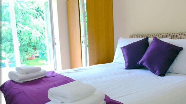 A neatly made bed with white linens and purple pillows, a purple bedspread, and folded white towels on the side. - Ivy Garden Cottage (Alexandria)