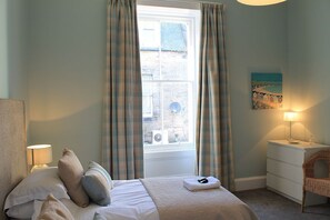 A bedroom with a bed, a bedside lamp, a nightstand, a chair, and a window with curtains.