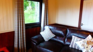A dark brown leather sofa with plaid cushions, a window with plaid curtains, and a door in a room with wooden paneling and a white wall. - Birch 22 With Hot Tub, Newton Stewart (Newton Stewart)