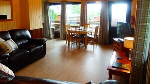 A living room with a wooden ceiling, a black leather sofa, a dining table with chairs, and a television. - Birch 22 With Hot Tub, Newton Stewart (Newton Stewart)
