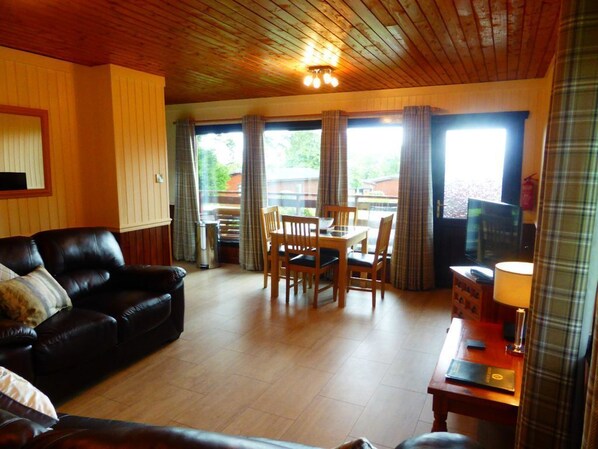 A living room with a wooden ceiling, a black leather sofa, a dining table with chairs, and a television.