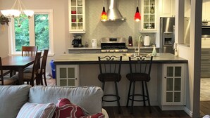 A modern kitchen with white cabinets, a stainless steel refrigerator, and a gas stove. There are two bar stools and a chandelier with red lampshades. - TO Stays - Waterfront Lake House Retreat (Ryerson)