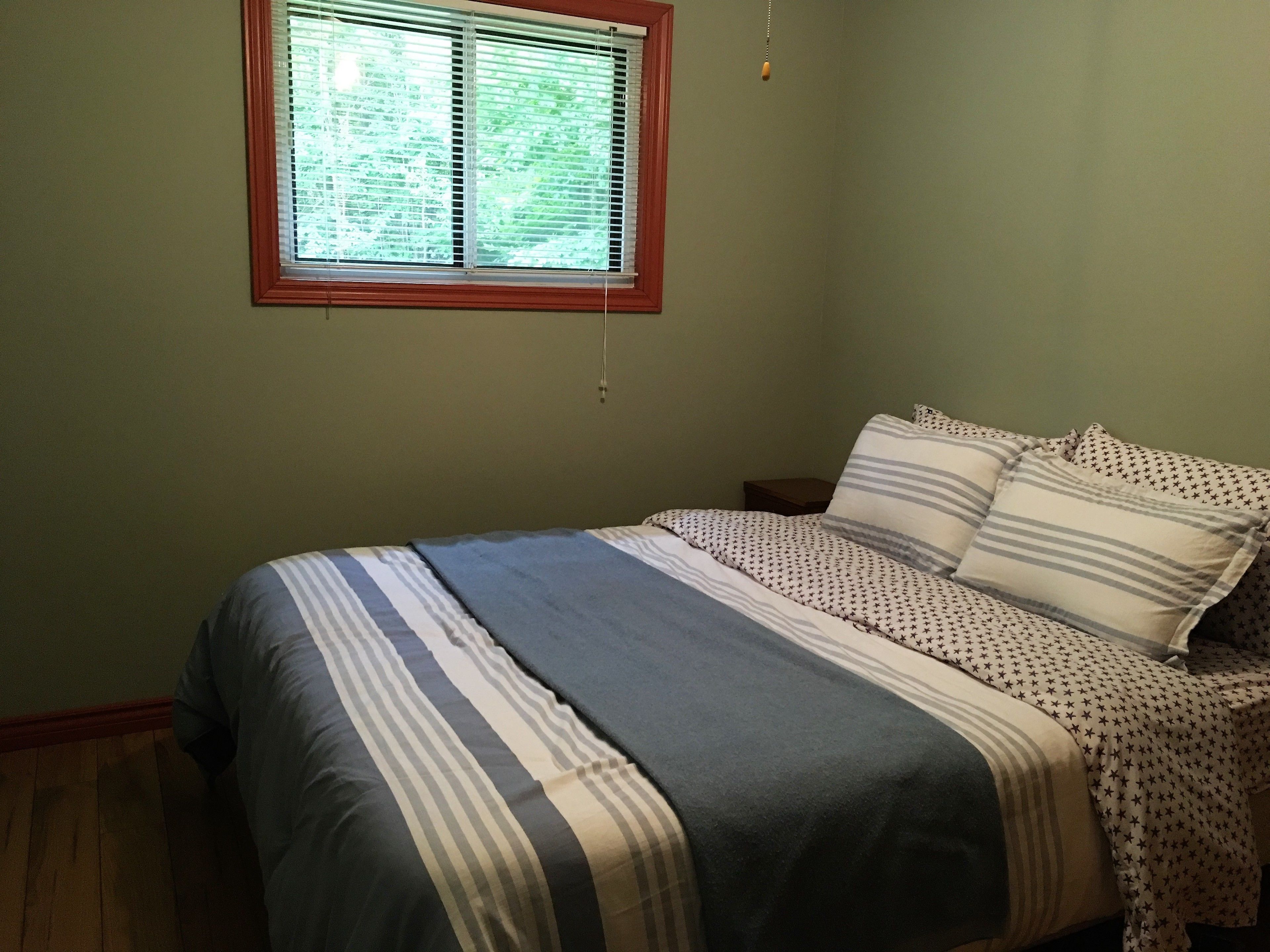 A bedroom with a bed, a window with blinds, a nightstand, and a hanging light fixture.