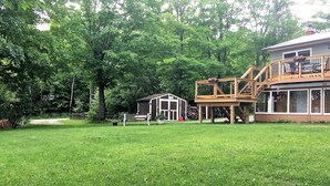 A large house with a wooden deck and a small shed in a grassy yard surrounded by trees. - TO Stays - Waterfront Lake House Retreat (Ryerson)