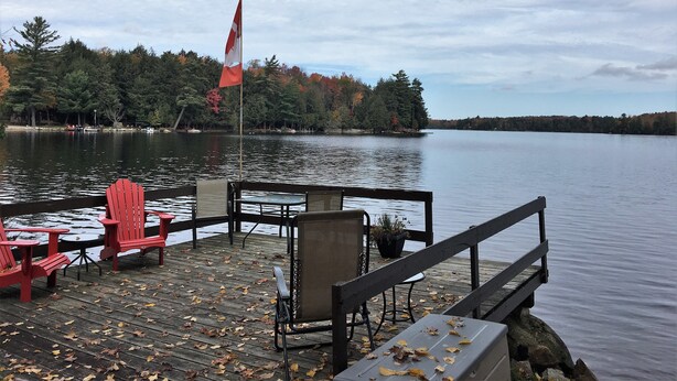 A wooden dock with red Adirondack chairs, a flag, and a calm lake with trees in the background. - TO Stays - Waterfront Lake House Retreat (Ryerson)