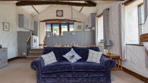 A cozy living room with a dark blue velvet sofa, a wooden staircase, and a view of the kitchen in the background. - Mill Barn (Hot Tub) 2 bedrooms (Brampton)