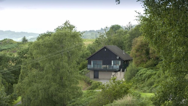A house with a balcony, surrounded by dense greenery and hills in the background. - Achmony Green lodge (Inverness)