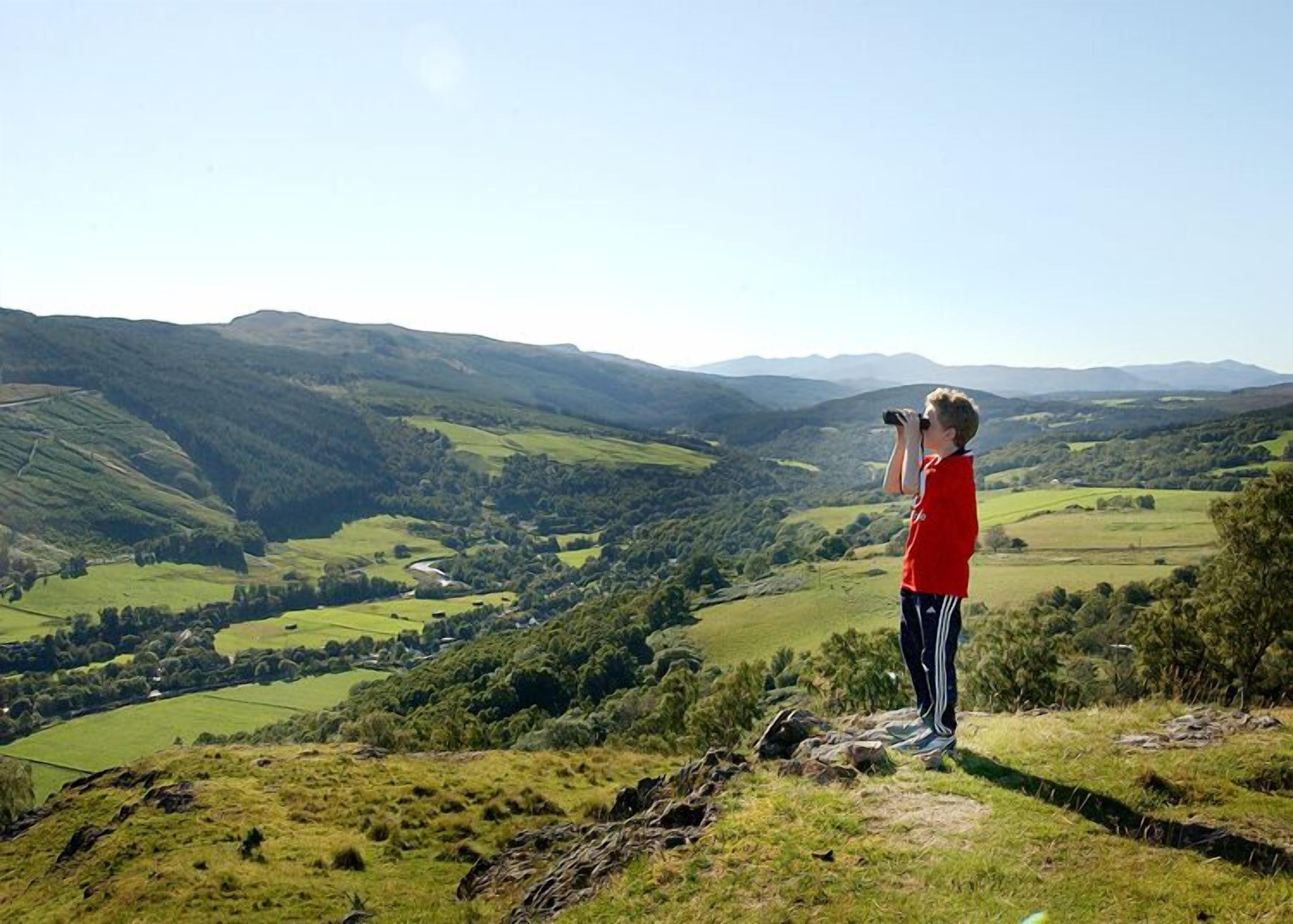 Una persona está de pie en una colina cubierta de hierba, contemplando un valle con colinas ondulantes y un río.