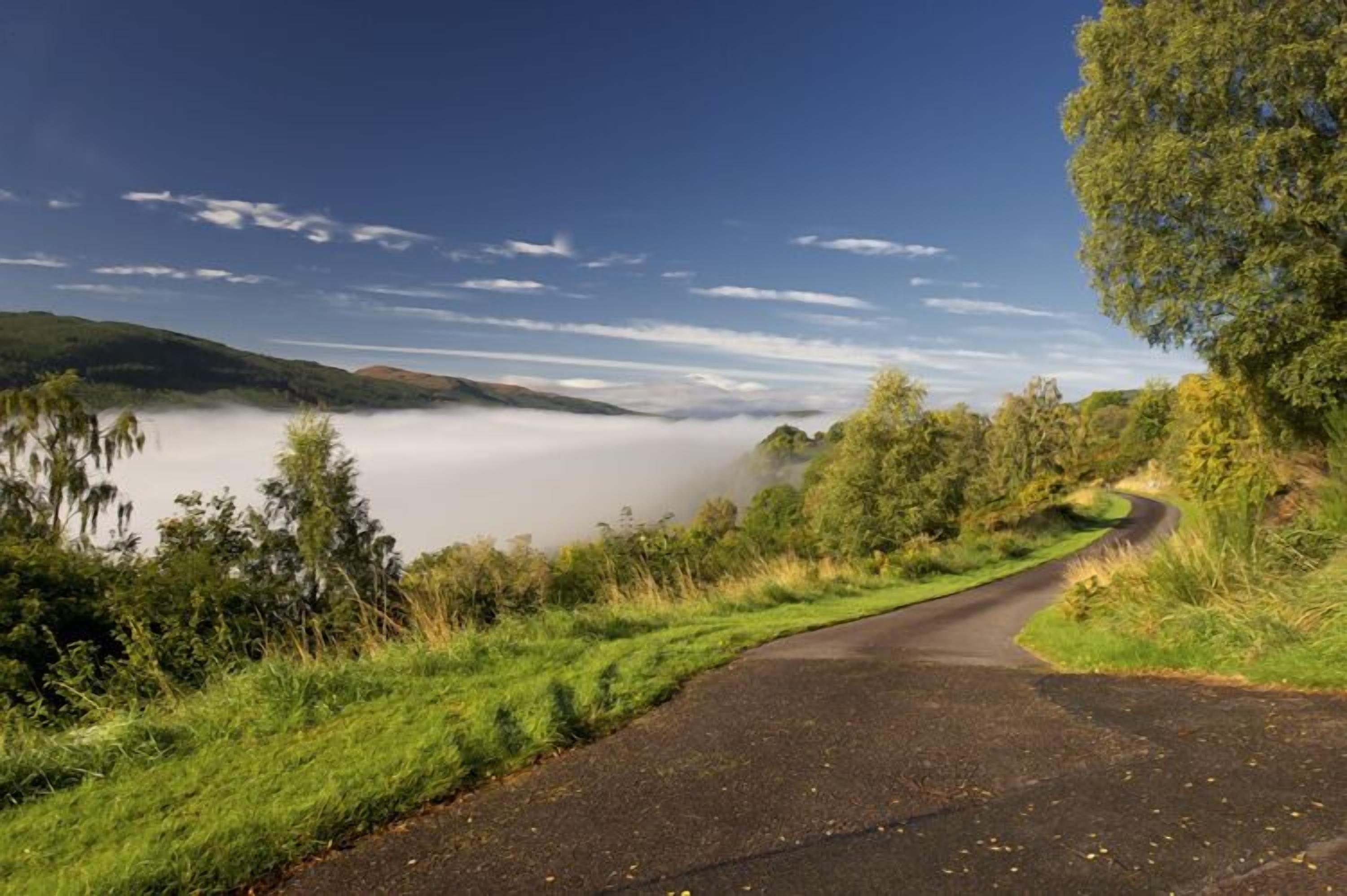 Eine kurvenreiche Straße schlängelt sich durch eine üppige Landschaft, mit einem See und nebelverhangenen Hügeln im Hintergrund.