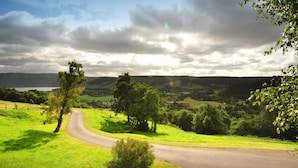 A rural road curving through a green landscape with trees and hills in the background. - Achmony Green lodge (Inverness)
