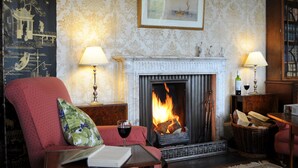 Un ambiente accogliente con camino, una poltrona rossa, un tavolo con un libro e un bicchiere di vino, e due lampade.