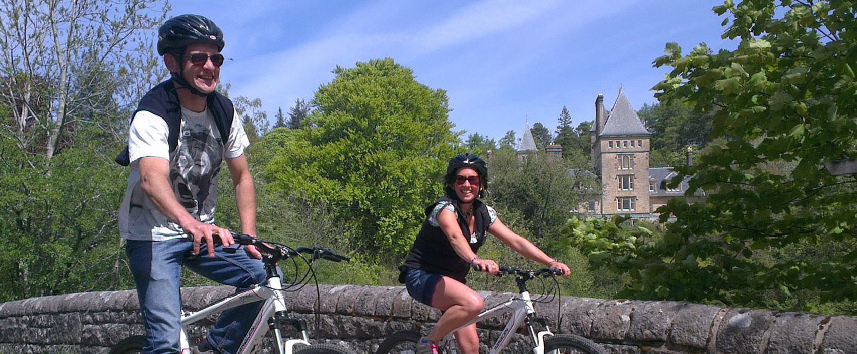 Two people on bicycles near a stone bridge with a historic building in the background.
