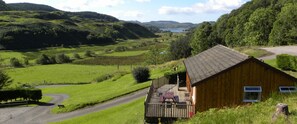 A wooden cabin with a porch, surrounded by lush green fields and hills. - Conifer Lodge (Oban)