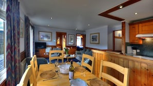 A dining area with a wooden table set for four, surrounded by chairs. There are placemats, cutlery, and a bottle of wine on the table. The room has wooden walls and ceiling, and there are framed pictures on the walls.