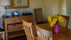 Un coin repas avec une table en bois, des chaises, un vase de fleurs jaunes, une lampe et une radio posée sur un meuble en bois.